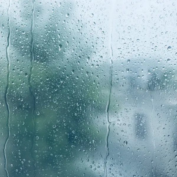 A close-up image of a foggy window with visible water droplets on the interior glass surface, illustrating condensation due to high humidity indoors.