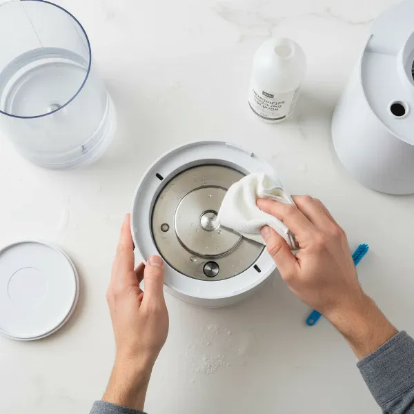 A person carefully cleaning the base of an ultrasonic humidifier with a cloth and a descaling solution, focusing on the transducer.